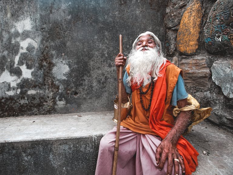 Portrait of an elderly Hindu man in vibrant traditional clothing, sitting with a walking stick.